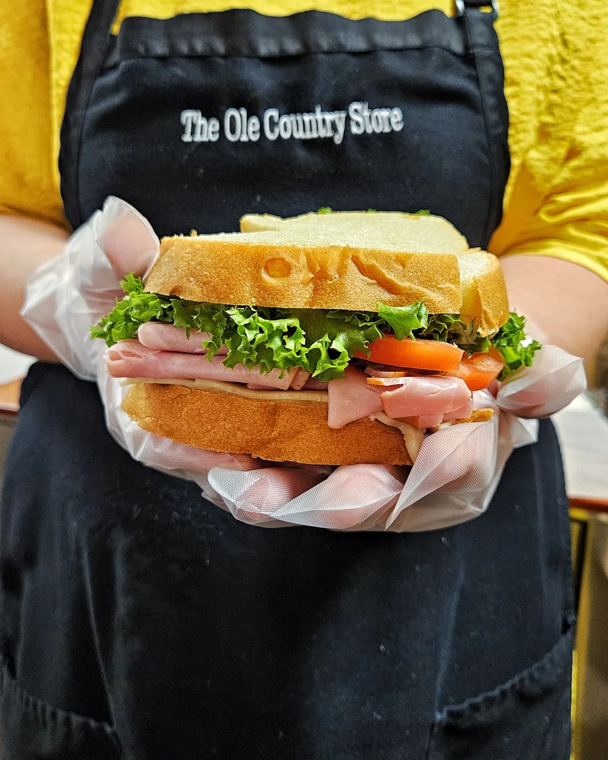 A deli worker in a Ole Country Store apron holding a made-to-order sandwich